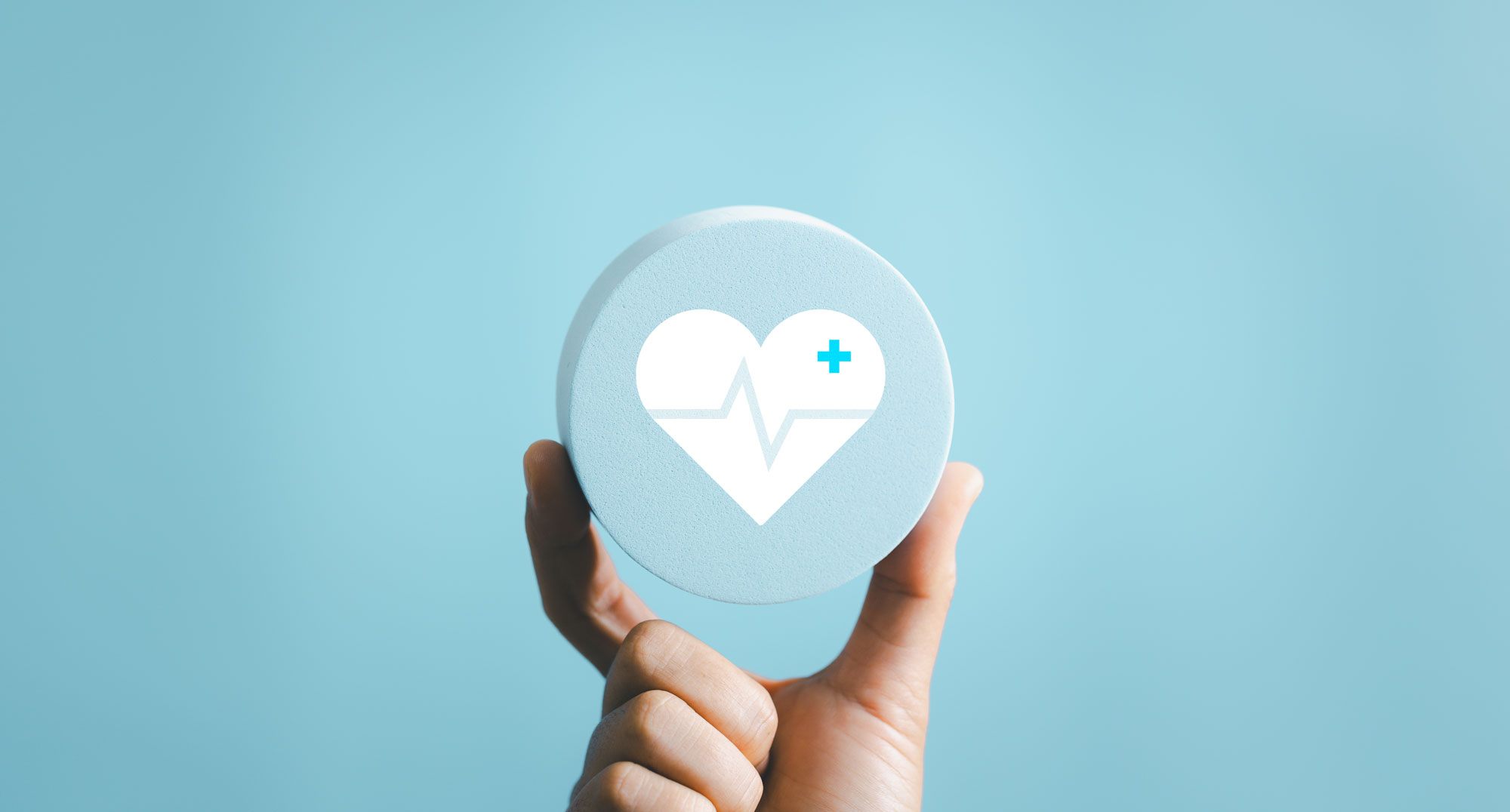 Eine Hand hält ein rundes Symbol mit einem Herz und medizinischem Pluszeichen auf hellblauem Hintergrund.